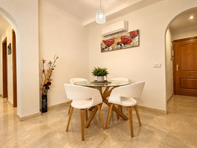 Zona de comedor compacta con mesa redonda de cristal y base de madera, cuatro sillas blancas y suelo de mármol claro. Un split de aire acondicionado y un cuadro floral decoran la pared.