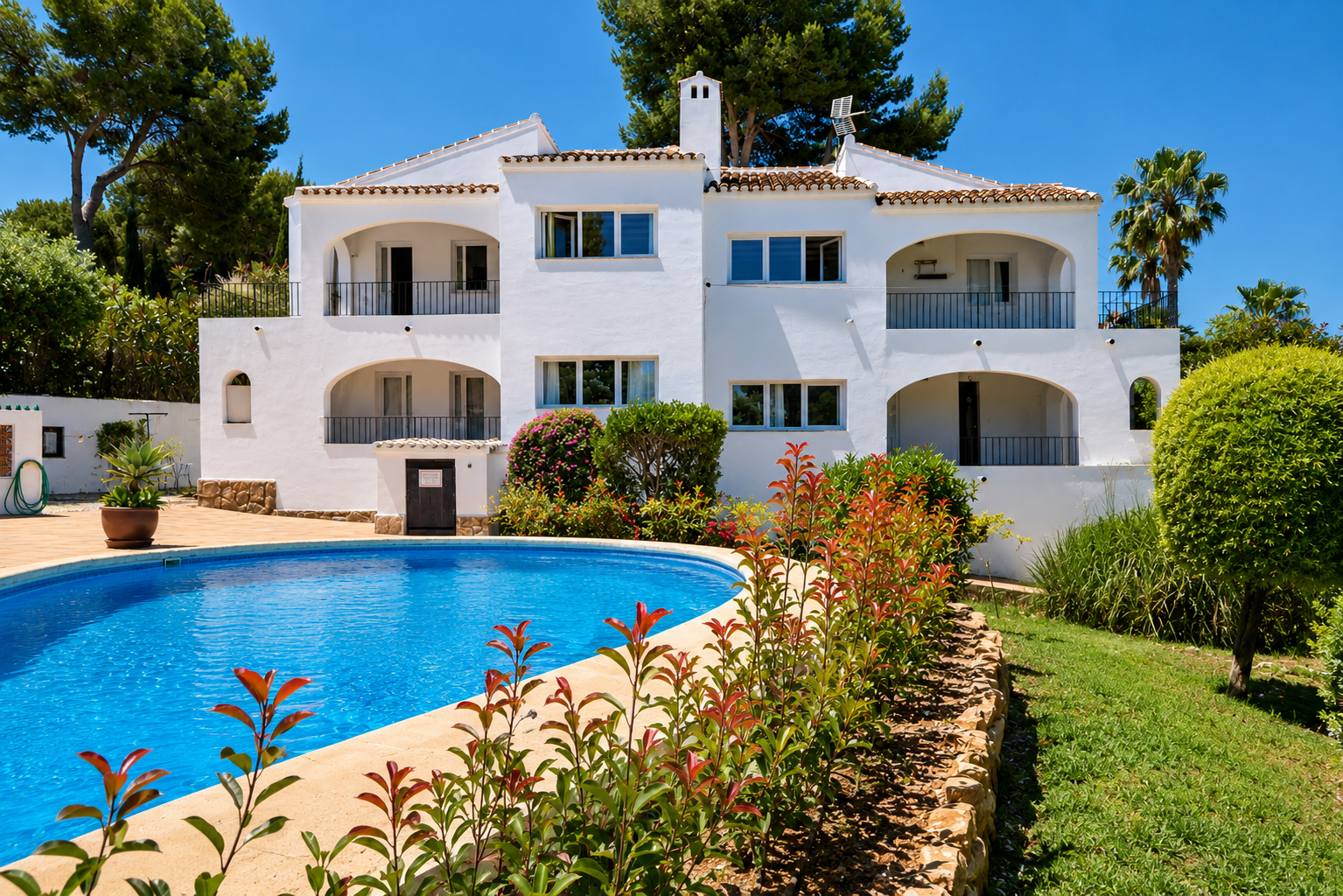 Edificio blanco de estilo mediterráneo con múltiples balcones arqueados y barandillas negras. La propiedad cuenta con una gran piscina curva y jardines exuberantes bajo un cielo azul.