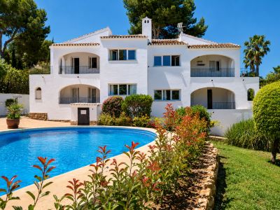 Edificio blanco de estilo mediterráneo con múltiples balcones arqueados y barandillas negras. La propiedad cuenta con una gran piscina curva y jardines exuberantes bajo un cielo azul.