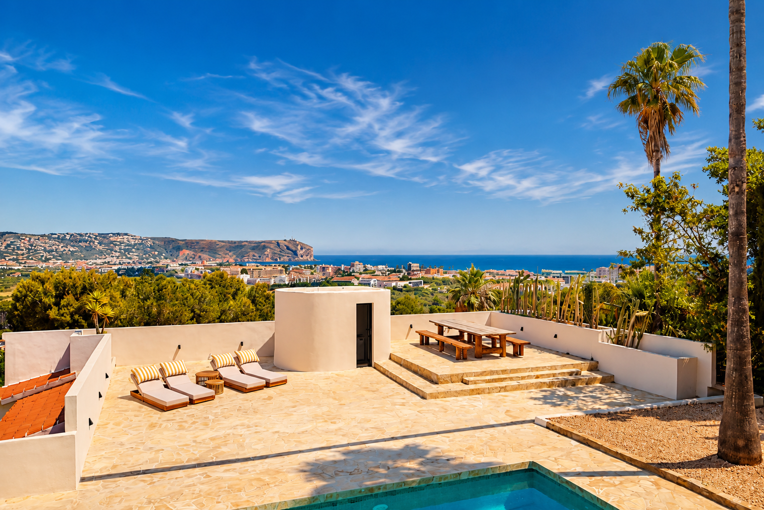 Amplia terraza de piedra con piscina, tumbonas y comedor de madera elevado, ofreciendo vistas panorámicas a la bahía de Jávea y la costa bajo un cielo azul.
