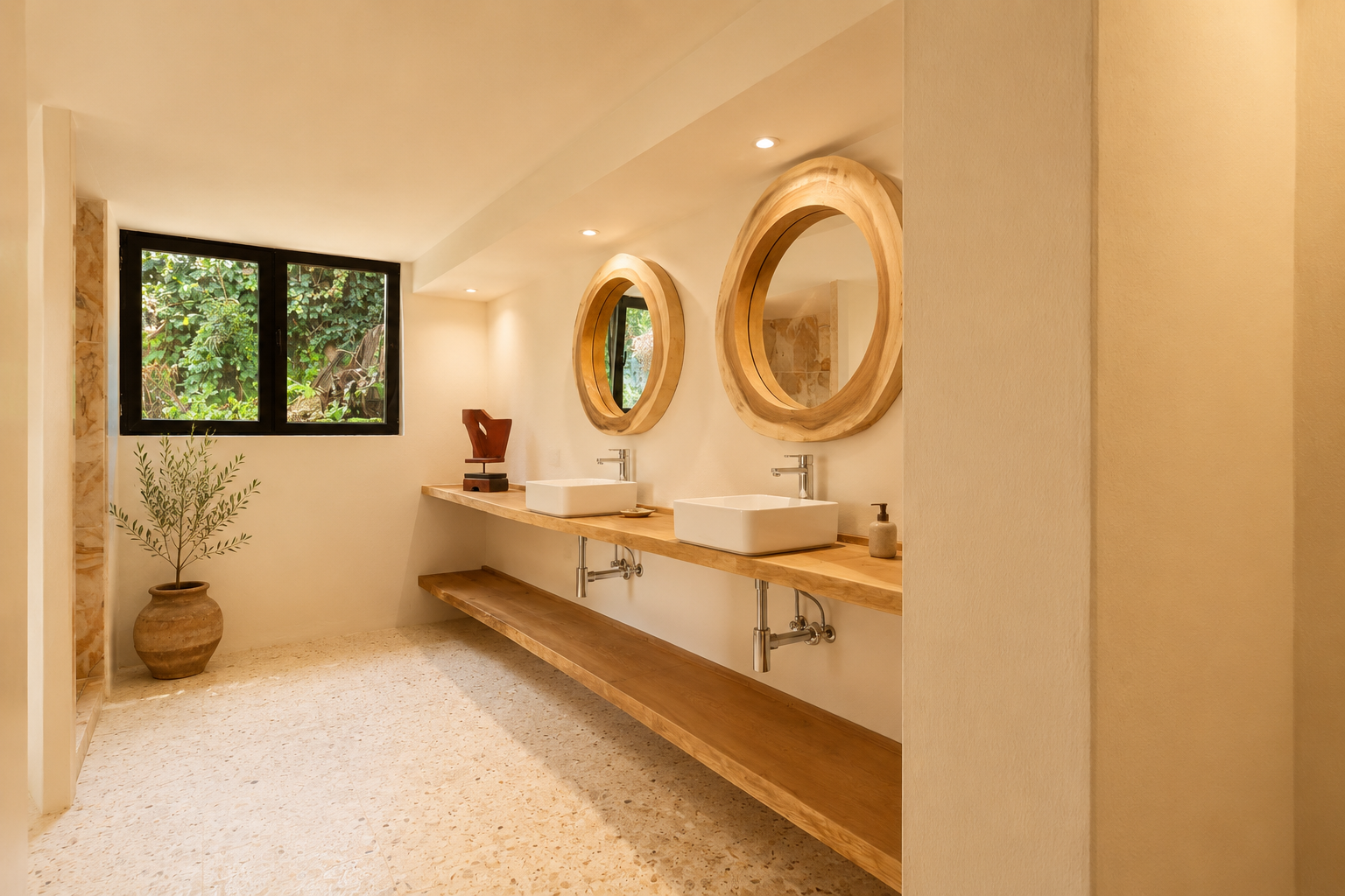 Baño con doble tocador de madera, lavabos rectangulares blancos y grandes espejos redondos con marcos de madera natural. Una ventana ofrece vistas a la vegetación, complementando el suelo de terrazo claro.