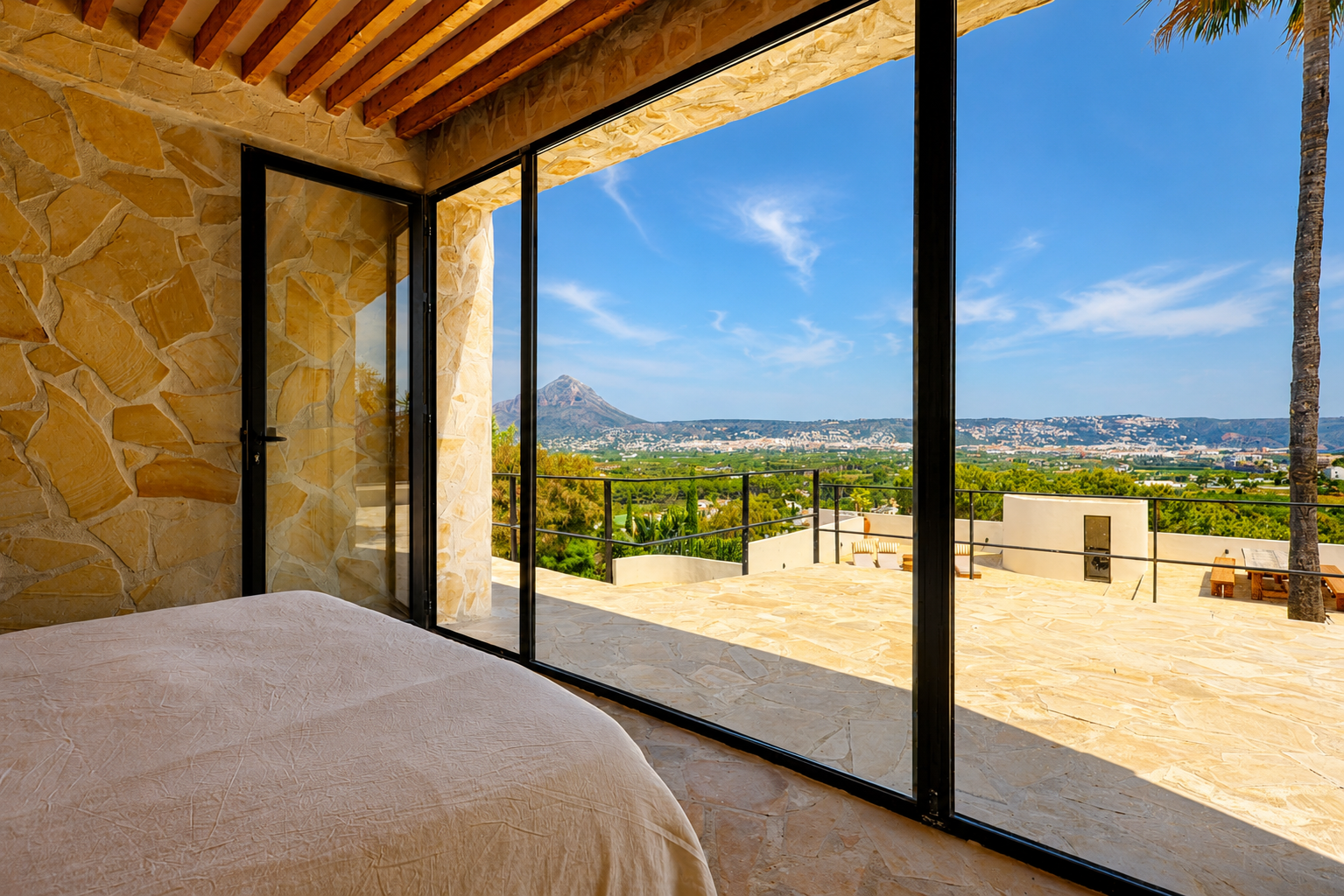 Dormitorio con paredes de piedra natural y vigas de madera, abriendo a una terraza con vistas panorámicas al Montgó y la bahía de Jávea.