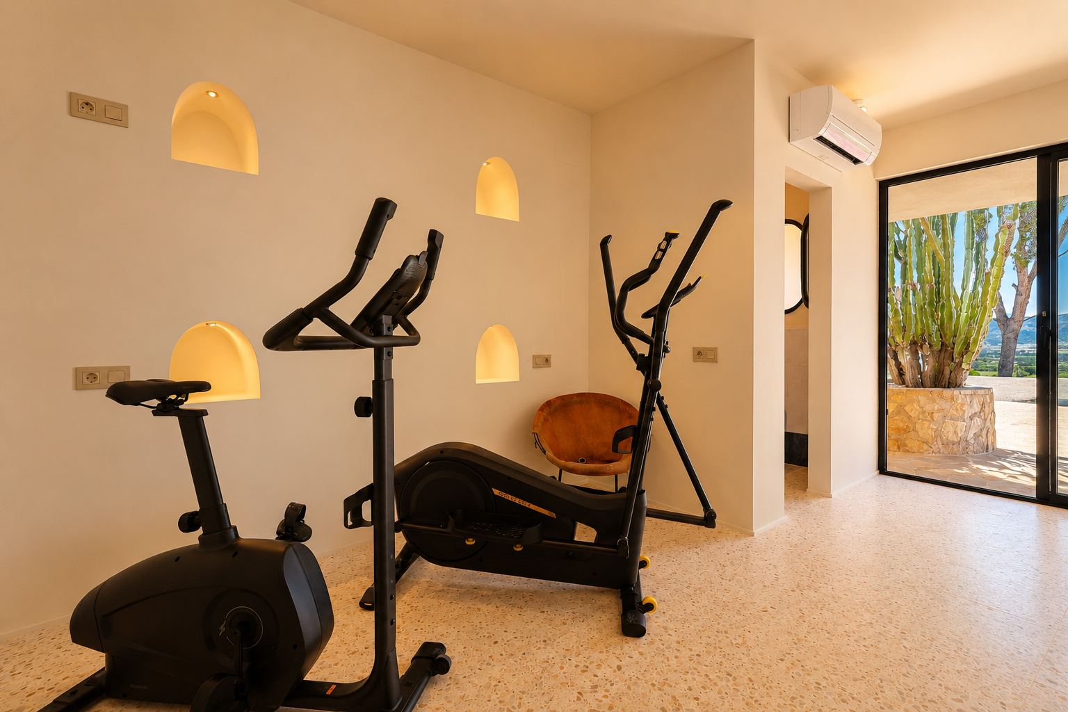 Espacio fitness equipado con bicicleta estática y elíptica, destacando nichos de pared iluminados y vistas a un jardín de cactus y montañas.