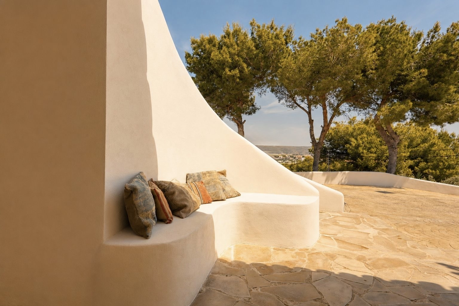 Esta terraza exterior presenta un banco curvo de estuco integrado con cojines bohemios, sobre un patio de losas irregulares. Pinos maduros y vistas lejanas al paisaje bajo un cielo despejado.