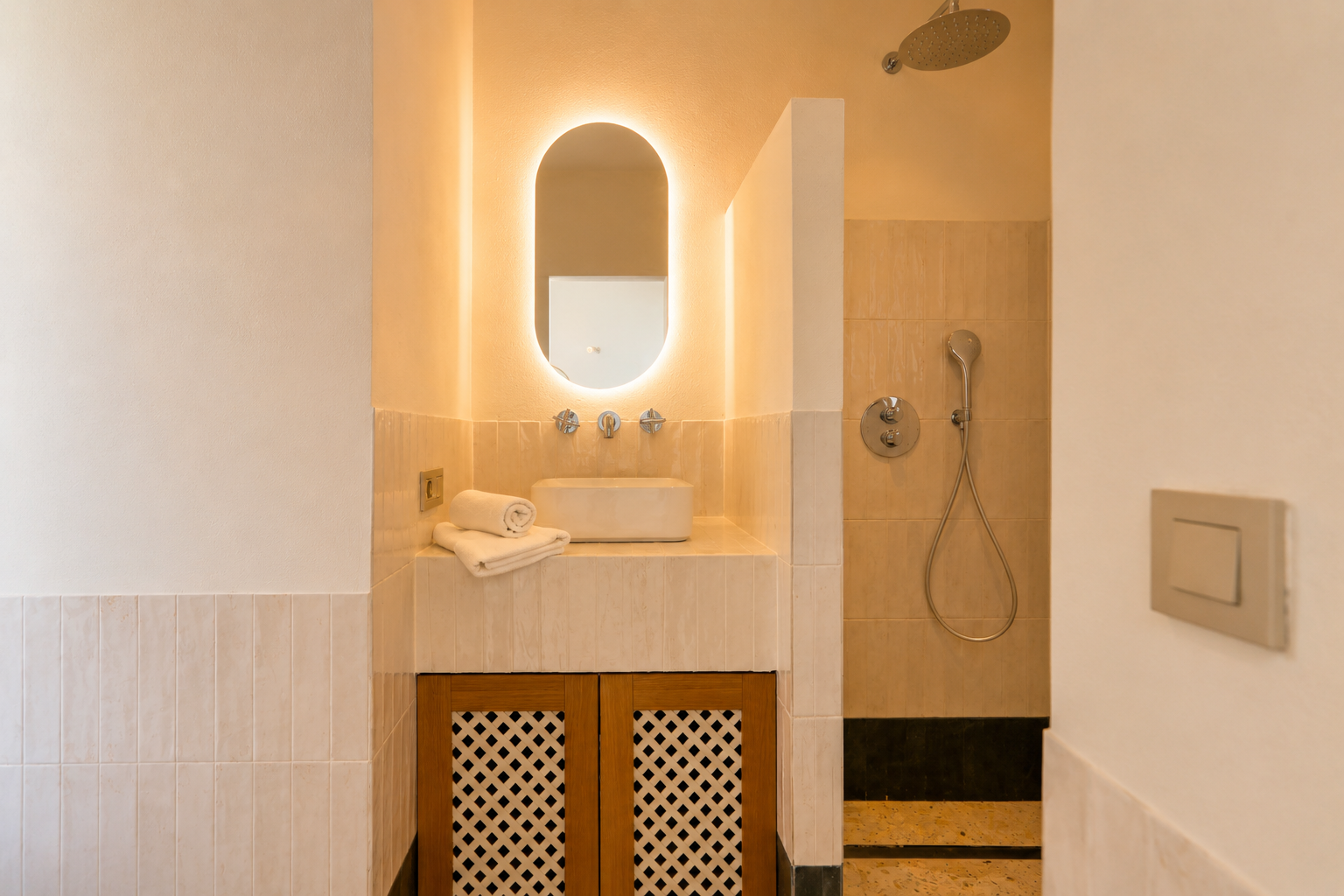 Este elegante baño presenta un lavabo sobre encimera, un espejo ovalado retroiluminado y un mueble con celosía de madera. La ducha contigua incluye un rociador de lluvia.