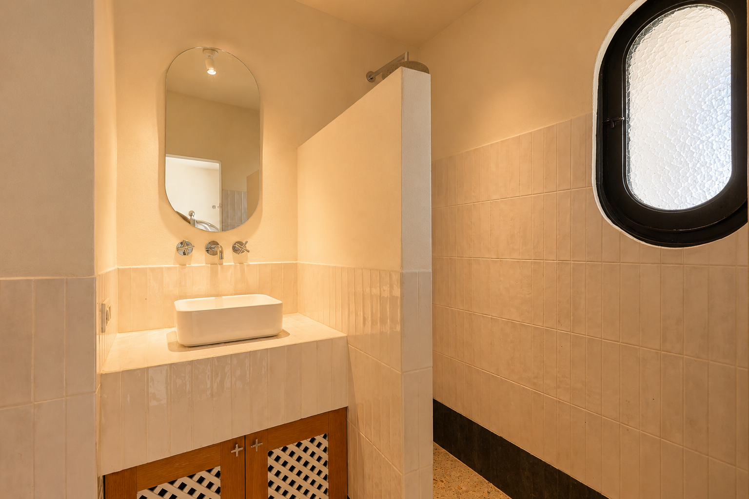 Lavabo de baño con lavamanos rectangular sobre encimera, grifería empotrada y espejo ovalado. Zona de ducha contigua con ventana ovalada esmerilada y paredes alicatadas en tonos claros.