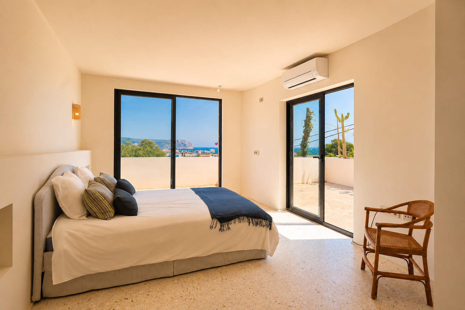 Luminoso dormitorio con suelo de terrazo claro y grandes puertas correderas de cristal que dan a una terraza privada. Ofrece amplias vistas al mar y la costa lejana.
