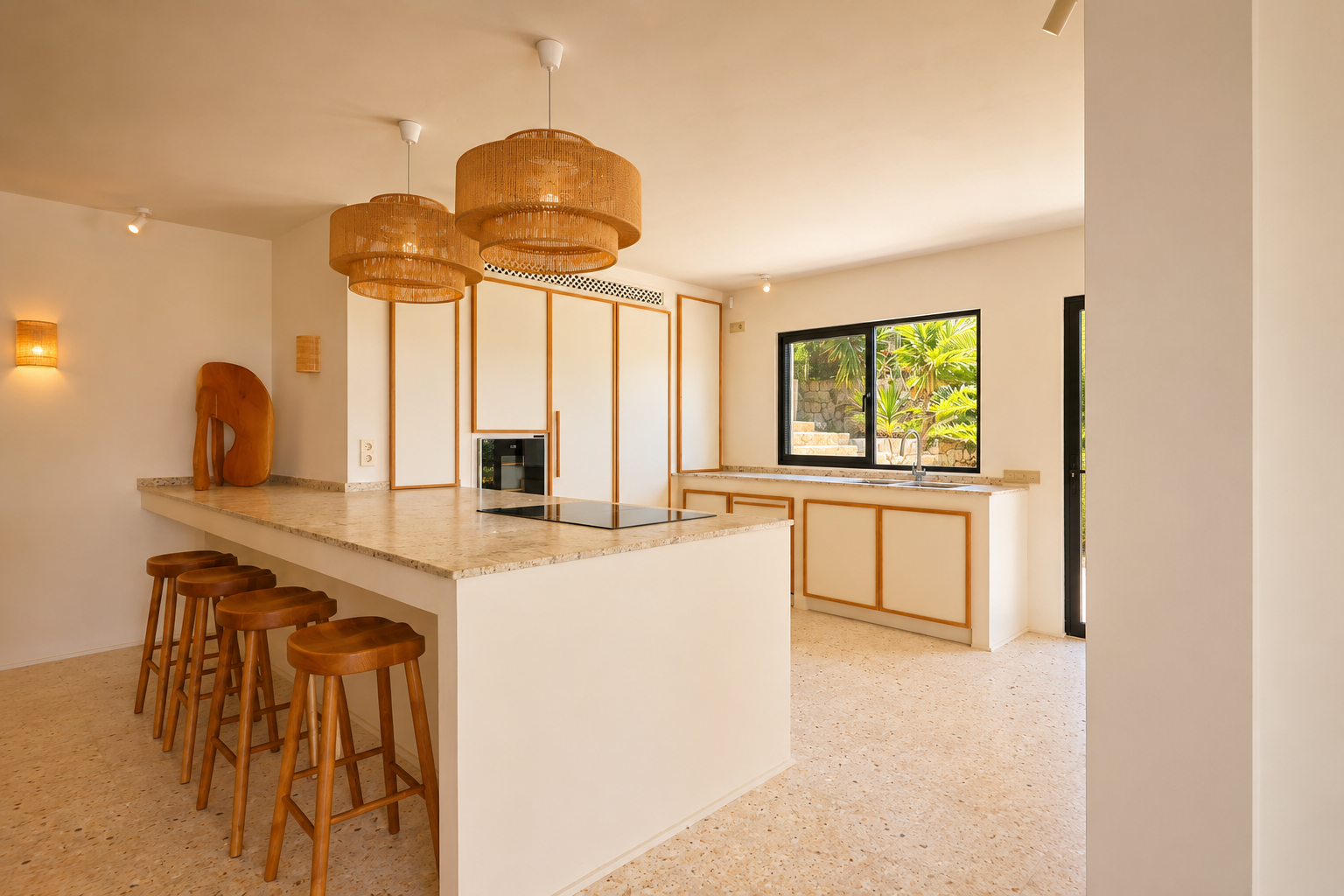 Moderna isla de cocina con placa integrada, encimera clara moteada y cinco taburetes de madera. Lámparas colgantes de mimbre iluminan el espacio, con vistas a la vegetación exterior.