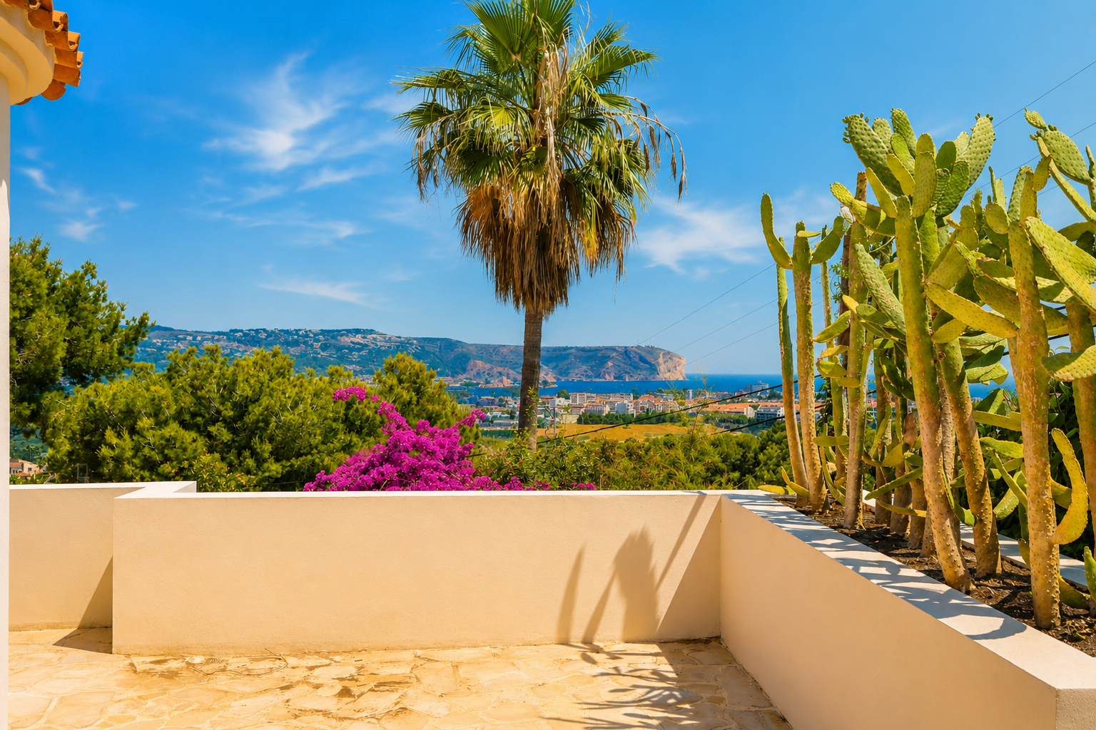 Terraza con suelo de piedra irregular y muro bajo color beige. Incluye un jardín de cactus y vistas panorámicas al mar Mediterráneo, la costa y un cabo lejano bajo cielo azul.