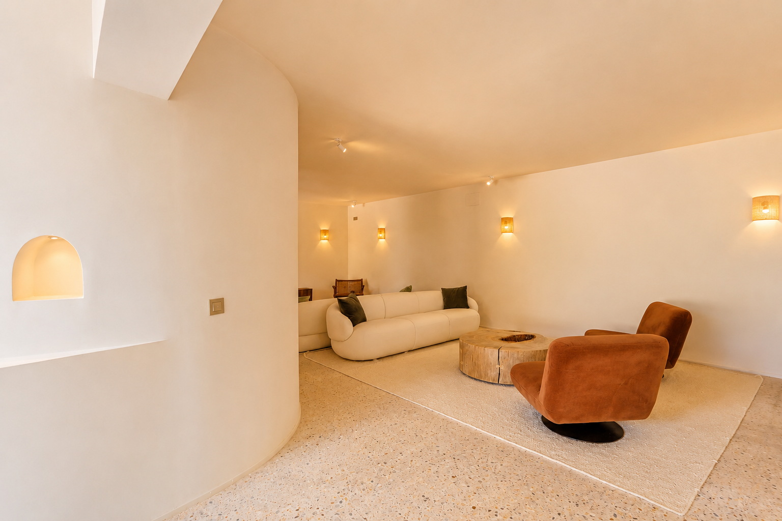 Zona de estar contemporánea con sofá curvo blanco roto, dos butacas color óxido y mesa de centro de madera rústica sobre suelo de terrazo claro. Apliques de pared tejidos ofrecen una iluminación cálida.