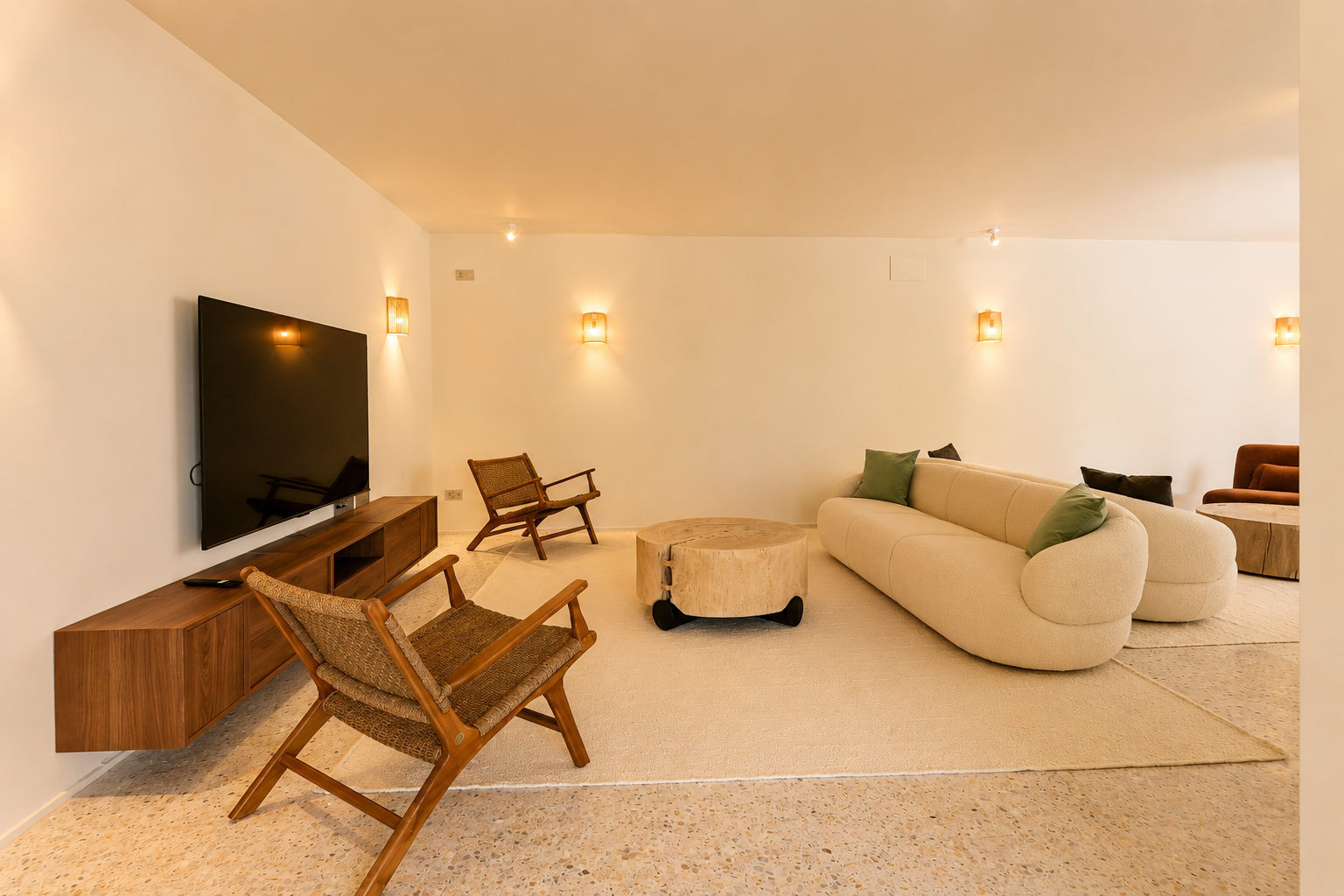 Zona de estar contemporánea con sofá curvo color crema, mesa de centro rústica de madera y sillones de mimbre sobre suelo de terrazo, iluminada por apliques cálidos.