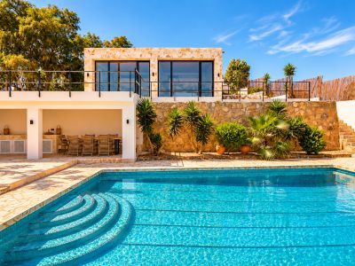 Amplia piscina exterior con escalones curvos, junto a una terraza pavimentada que conecta con un comedor cubierto y cocina exterior. Arriba, edificio de piedra con balcón y puertas de cristal.