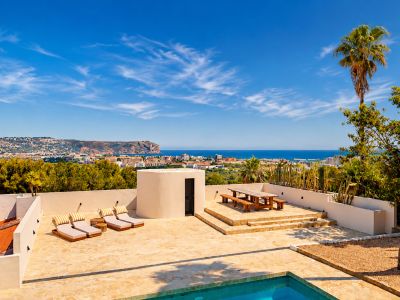 Amplia terraza de piedra con piscina, tumbonas y comedor de madera elevado, ofreciendo vistas panorámicas a la bahía de Jávea y la costa bajo un cielo azul.