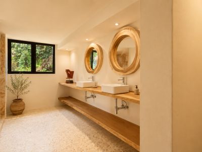 Baño con doble tocador de madera, lavabos rectangulares blancos y grandes espejos redondos con marcos de madera natural. Una ventana ofrece vistas a la vegetación, complementando el suelo de terrazo claro.