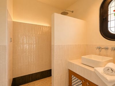Baño moderno con ducha a ras de suelo, rociador de lluvia y luz LED indirecta. Lavabo sobre encimera de azulejos y una distintiva ventana ovalada de vidrieras con motivo de rosa.