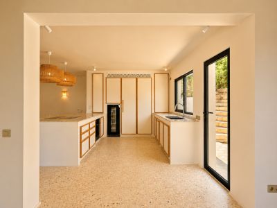 Cocina moderna con armarios de ribete de madera clara, suelo tipo terrazo y una gran isla con placa integrada. Lámparas colgantes de rafia iluminan el espacio.