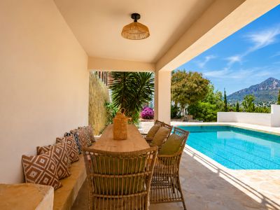 Comedor exterior cubierto con mesa de madera y asientos de mimbre, adyacente a la piscina. Ofrece vistas al Montgó y vegetación mediterránea sobre suelo de piedra.