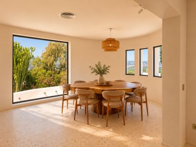 Comedor luminoso con mesa de madera orgánica para ocho personas y suelo de terrazo. Grandes ventanales curvos ofrecen vistas a la vegetación exuberante y al paisaje lejano.