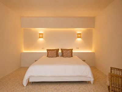 Dormitorio con cama doble, iluminación LED integrada en el cabecero y apliques de pared de rafia. Suelo de terrazo y cojines decorativos.
