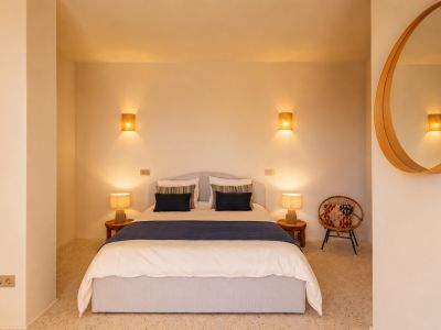 Dormitorio con cama doble, mesitas de noche de madera y lámparas de mesa. Destacan apliques de pared con pantallas de rafia y un espejo redondo de madera. Suelo de terrazo.