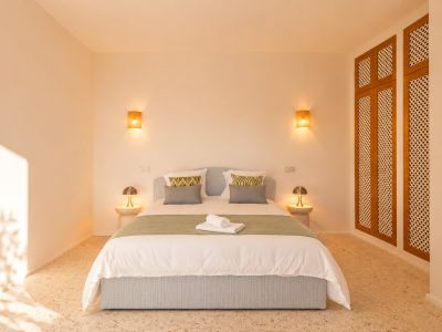 Dormitorio con cama tapizada en azul claro, suelo de terrazo y luz cálida de apliques de pared tejidos. Armario empotrado de madera con celosía a la derecha.