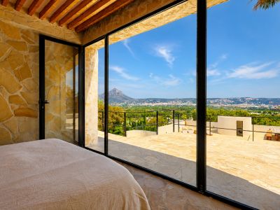 Dormitorio con paredes de piedra natural y vigas de madera, abriendo a una terraza con vistas panorámicas al Montgó y la bahía de Jávea.