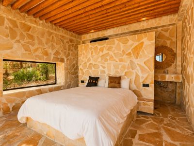 Dormitorio con paredes y suelo de piedra natural irregular, techo de vigas de madera y ventana horizontal con vistas a la vegetación. Incluye un lavabo integrado con espejo de ratán.