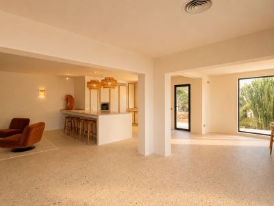 Espacioso salón-comedor de concepto abierto con suelo de terrazo, integrando una cocina con isla y un rincón de estar, iluminado por luz natural y vistas a la exuberante vegetación exterior.