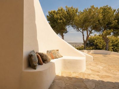 Esta terraza exterior presenta un banco curvo de estuco integrado con cojines bohemios, sobre un patio de losas irregulares. Pinos maduros y vistas lejanas al paisaje bajo un cielo despejado.