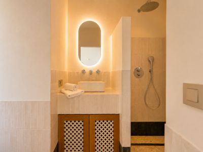 Este elegante baño presenta un lavabo sobre encimera, un espejo ovalado retroiluminado y un mueble con celosía de madera. La ducha contigua incluye un rociador de lluvia.