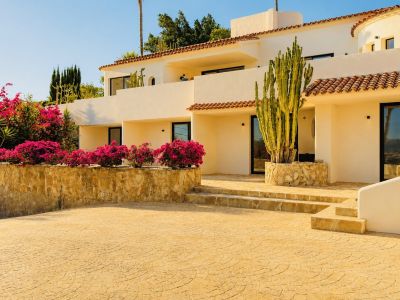 Fachada mediterránea color crema con tejas de terracota, enmarcada por buganvillas fucsia y un cactus alto, con acceso pavimentado y escalones de piedra.