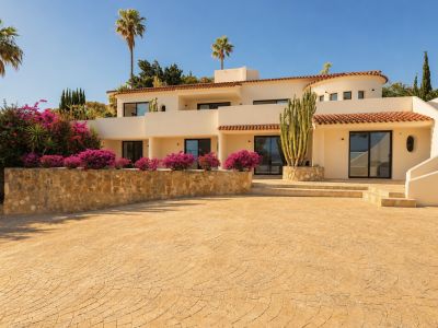 La fachada color crema de la villa, tejas de terracota y grandes puertas de cristal, enmarcada por buganvillas vibrantes, un cactus alto y palmeras bajo un cielo azul. Un camino estampado conduce a la entrada.