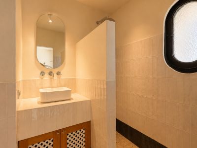 Lavabo de baño con lavamanos rectangular sobre encimera, grifería empotrada y espejo ovalado. Zona de ducha contigua con ventana ovalada esmerilada y paredes alicatadas en tonos claros.