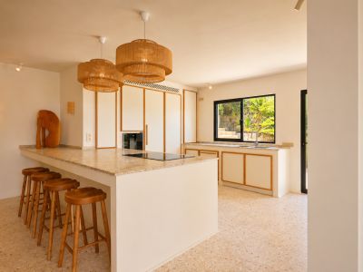 Moderna isla de cocina con placa integrada, encimera clara moteada y cinco taburetes de madera. Lámparas colgantes de mimbre iluminan el espacio, con vistas a la vegetación exterior.