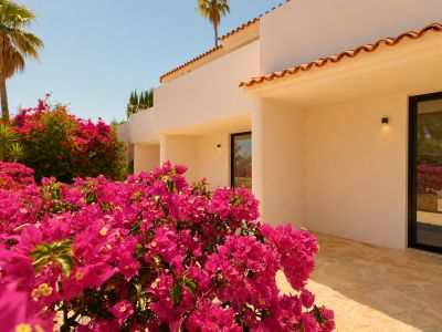 Primer plano de exuberantes buganvillas fucsias que realzan la fachada mediterránea en tonos claros, con tejas de barro y un sendero de piedra irregular. Las ventanas oscuras reflejan el entorno soleado.