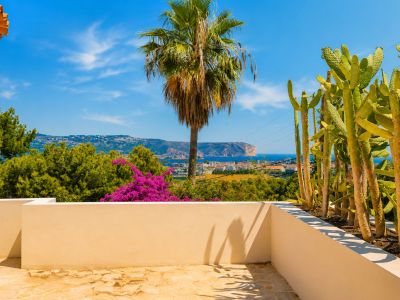 Terraza con suelo de piedra irregular y muro bajo color beige. Incluye un jardín de cactus y vistas panorámicas al mar Mediterráneo, la costa y un cabo lejano bajo cielo azul.