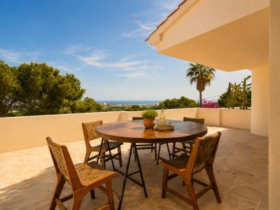 Terraza exterior con mesa redonda de madera y sillas de mimbre sobre suelo de losas, ofreciendo vistas panorámicas al mar y la costa.