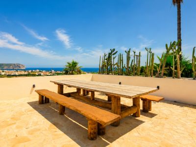 Terraza exterior con mesa y bancos de madera rústica, suelo de piedra clara y vistas panorámicas al mar y la ciudad, enmarcada por cactus.