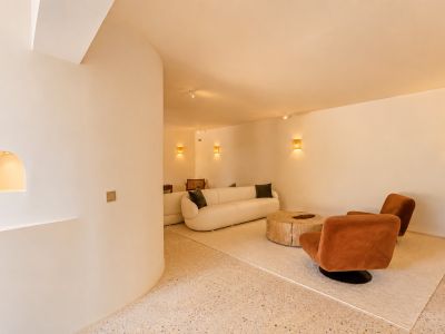 Zona de estar contemporánea con sofá curvo blanco roto, dos butacas color óxido y mesa de centro de madera rústica sobre suelo de terrazo claro. Apliques de pared tejidos ofrecen una iluminación cálida.
