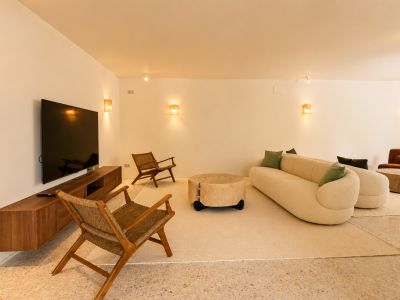 Zona de estar contemporánea con sofá curvo color crema, mesa de centro rústica de madera y sillones de mimbre sobre suelo de terrazo, iluminada por apliques cálidos.