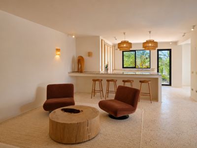 Zona de estar de concepto abierto con dos sillones color óxido, una mesa de centro rústica de madera y suelo de terrazo claro. El espacio conecta con una barra iluminada por lámparas de mimbre y vistas a la vegetación.