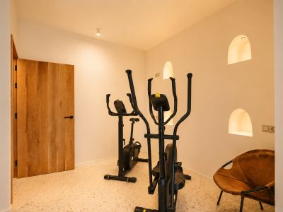 Zona de fitness dedicada con elíptica y bicicleta estática sobre suelo de terrazo, complementada por nichos arqueados iluminados y una puerta de madera.