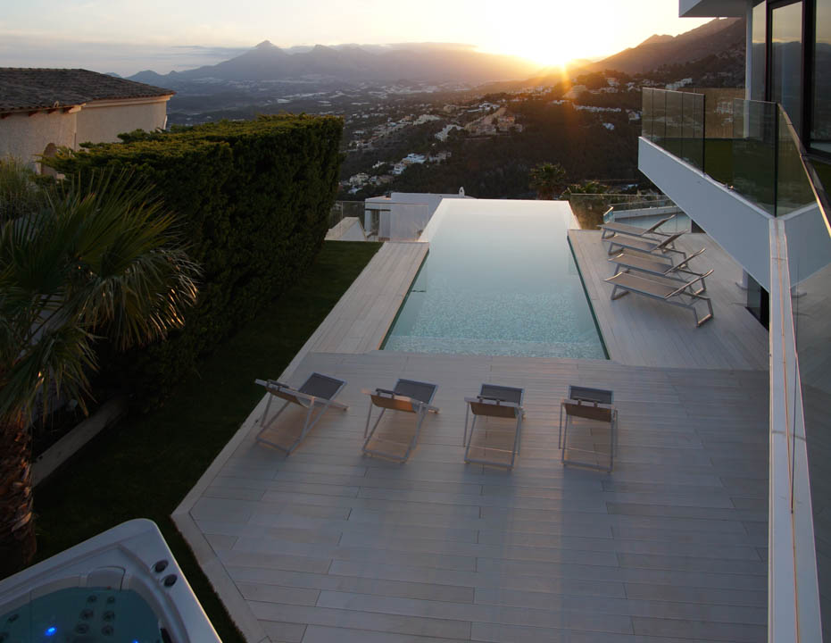 Amplia piscina infinita con tarima clara, varias tumbonas y jacuzzi separado, enmarcada por un paisaje montañoso con puesta de sol panorámica.