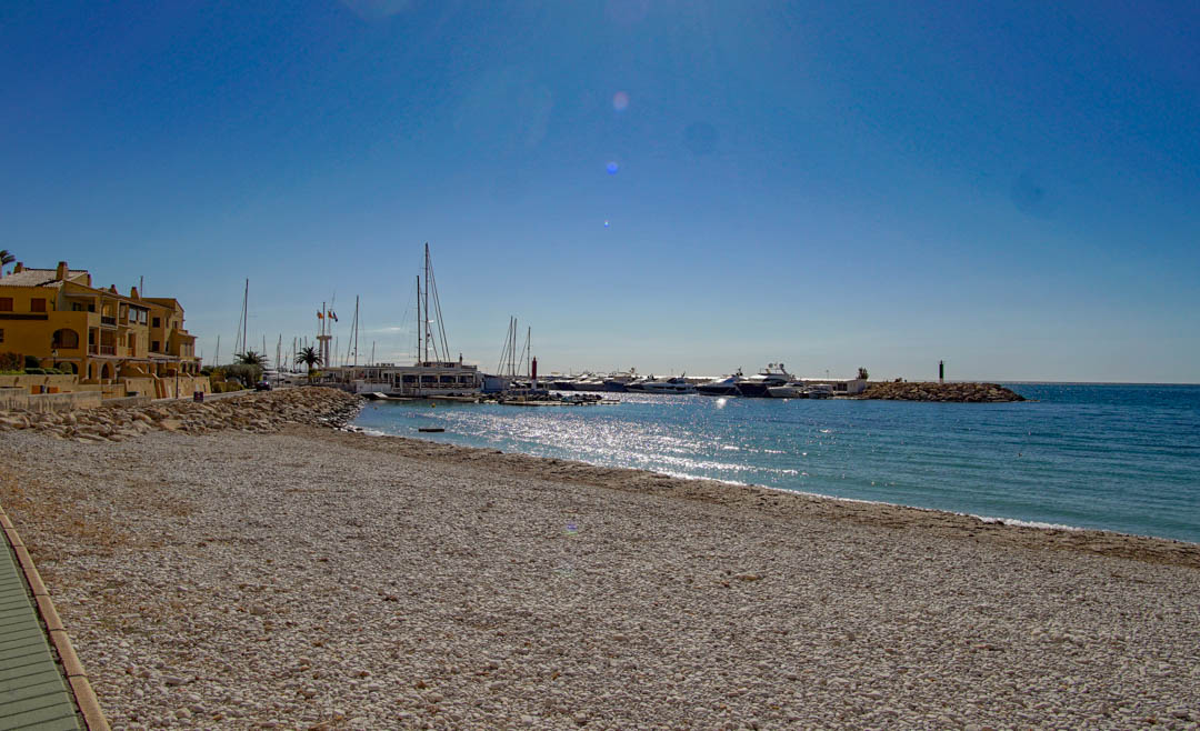 Amplia playa de guijarros se extiende hacia un puerto deportivo con yates, flanqueada por edificios costeros tradicionales bajo un cielo azul claro.