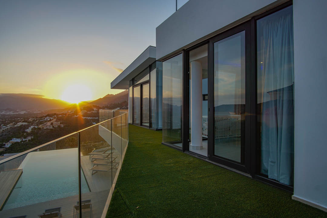 Amplia terraza con piscina infinita, tarima de madera y barandilla de cristal, ofreciendo vistas panorámicas al atardecer sobre montañas y valle.