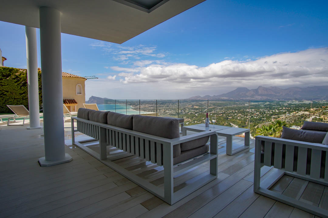 Amplia terraza cubierta con tarima clara y mobiliario lounge blanco, enmarcando una vista panorámica al mar, la costa y las montañas a través de una barandilla de cristal.