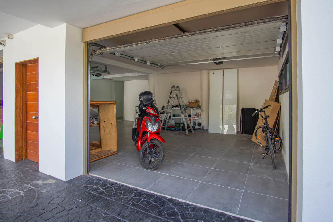 Amplio garaje con suelo de baldosas grises, puerta seccional parcialmente abierta y una puerta de entrada de madera a la izquierda. En el interior se ven una moto roja y una bicicleta.