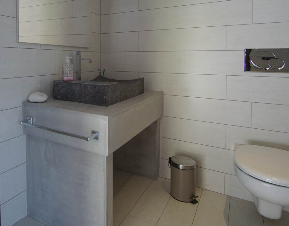 Aseo moderno con lavabo de piedra sobre encimera de hormigón y azulejos claros. Inodoro suspendido con pulsador cromado.