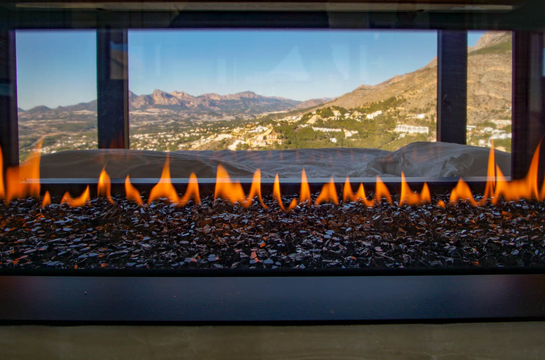 Chimenea de gas moderna con llamas sobre cristales oscuros, enmarcando una vista panorámica a montañas y un valle.