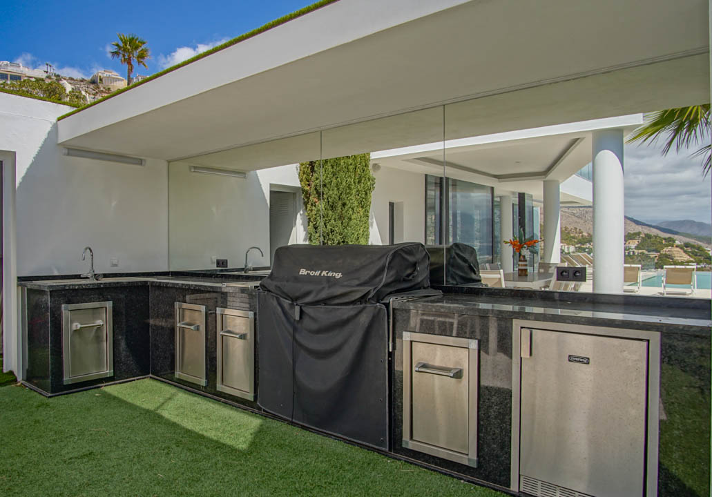 Cocina exterior en forma de L con encimeras de granito negro, fregaderos dobles, barbacoa cubierta y electrodomésticos de acero inoxidable, con vistas reflejadas a la piscina.